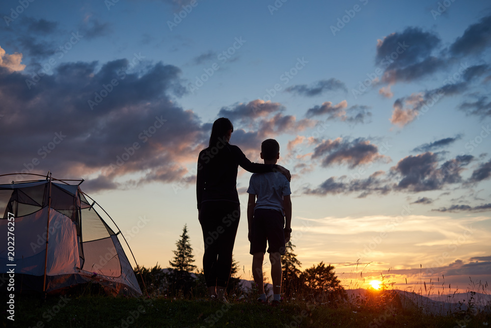Rear view of the mother hugs her son near the tent on top of the mountain enjoying the view of the sunset, on the background - mountains and hills under incredibly beautiful evening sky with clouds