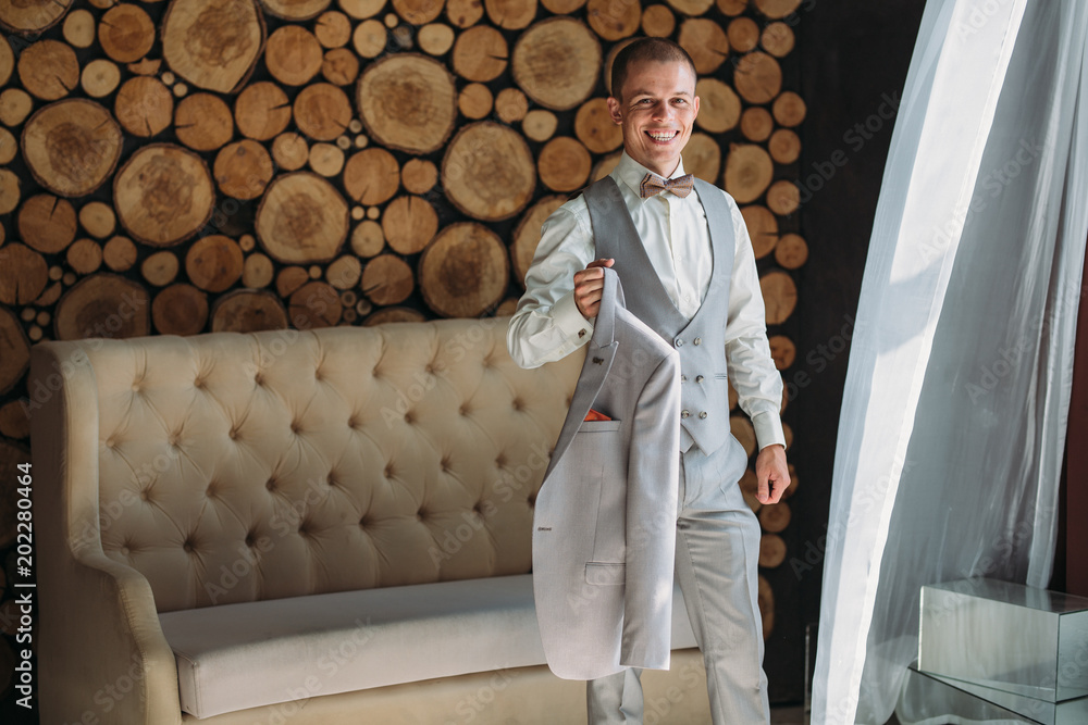 Fototapeta premium Preparation of the groom for the wedding day. Man dresses a white shirt in the morning in the studio with a wooden background. Blond businessman in strict business attire. Concept of business clothes