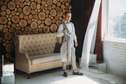 Preparation of the groom for the wedding day. Man dresses a white shirt in the morning in the studio with a wooden background. Blond businessman in strict business attire. Concept of business clothes © romannoru