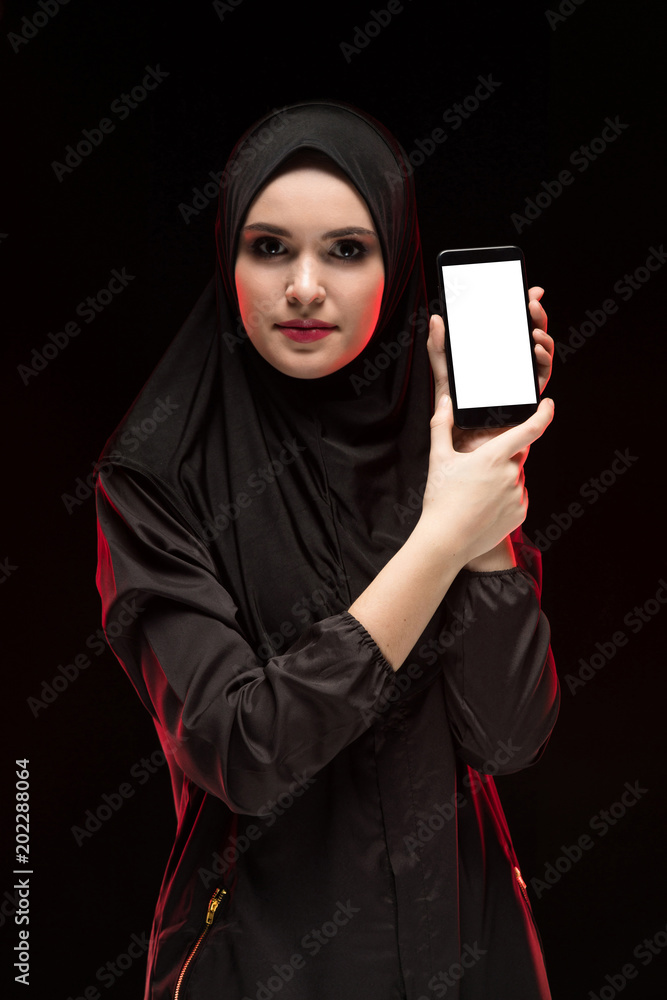 Portrait of beautiful smart young muslim woman wearing black hijab advertising mobile phone in her hands as education concept on black background Portrait of beautiful smart young muslim woman wearing black hijab advertising mobile phone in her hands as education concept on black background