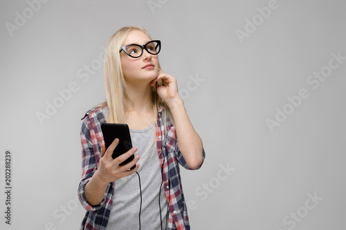 Portrait of attractive smiling sweet adorable blonde teenager girl in checkered clothes in glasses holding mobile phone in her hands in headphones on gray background