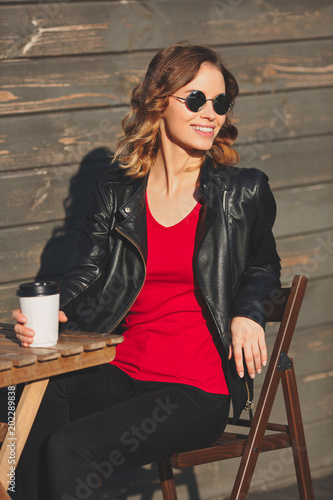 Young beautiful smiling woman in round glasses and in a black leather jacket drinking coffee