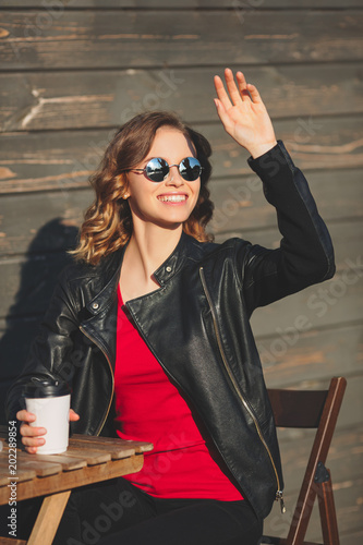 Young beautiful smiling woman in round glasses and in a black leather jacket drinking coffee