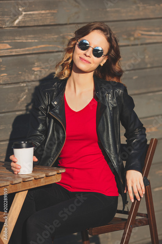 Young beautiful smiling woman in round glasses and in a black leather jacket drinking coffee