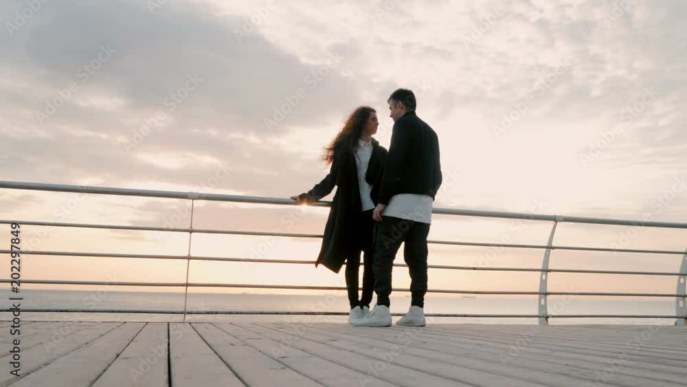 Young happy couple in love. Pair standing on wooden pier near sea or ocean and smiling to each other. People in black and white trendy clothing, They enjoying life. Romantic concept. 4k Young happy couple in love. Pair standing on wooden pier near sea or ocean and smiling to each other. People in black and white trendy clothing, They enjoying life. Romantic concept. 4k