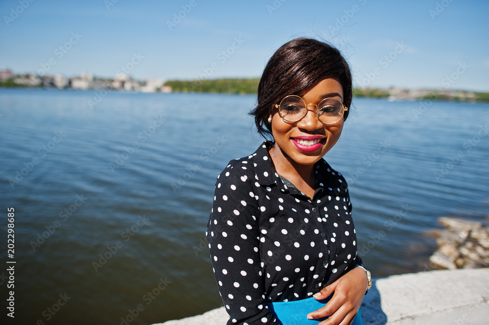 Stylish african american model girl in glasses, blue skirt and black blouse posed outdoor on bridge against lake. Stylish african american model girl in glasses, blue skirt and black blouse posed outdoor on bridge against lake.