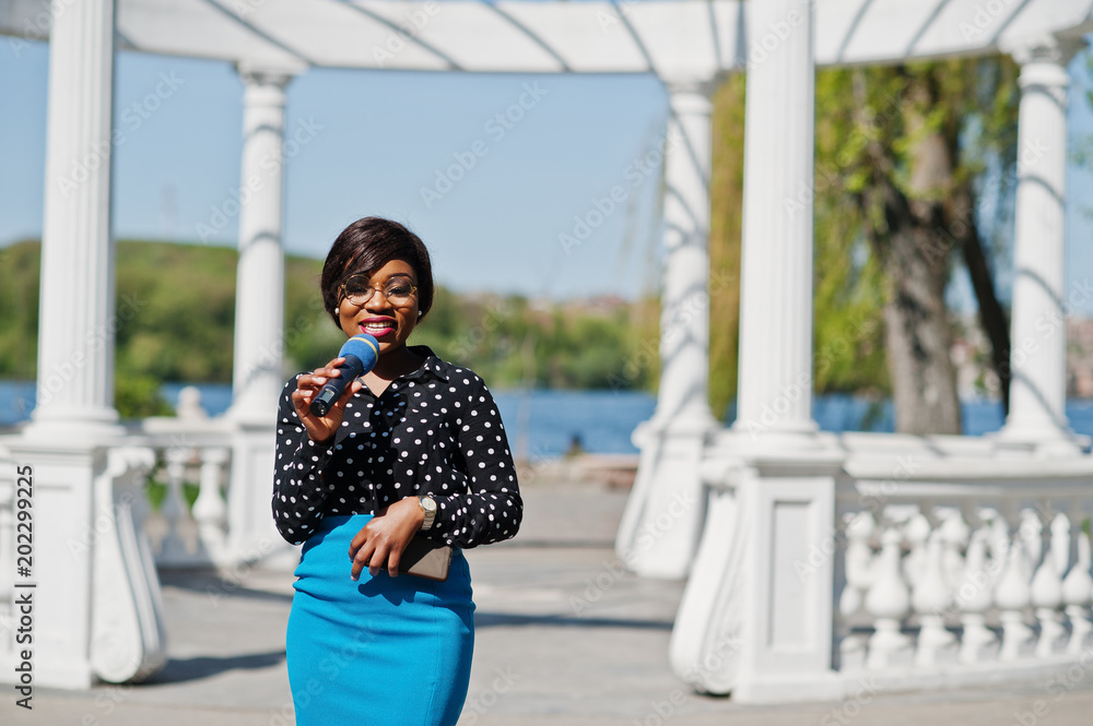 Stylish african american model girl TV presenter with microphone in glasses, blue skirt and black blouse posed outdoor against white stone arch with mobile phone.