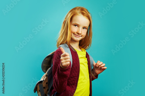 Φωτογραφία Positive schoolchild with backpack behind his shoulders shows his thumb, showing positive attitude to study, standing on blue isolated background