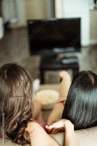 Back view photo of two girlfriends who are sitting on the couch watching TV.