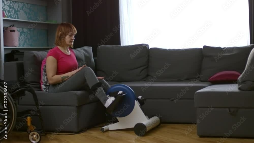 PAN of cheerful paraplegic woman sitting on coach and using rehabilitation cycling machine; wheelchair standing beside her PAN of cheerful paraplegic woman sitting on coach and using rehabilitation cycling machine; wheelchair standing beside her