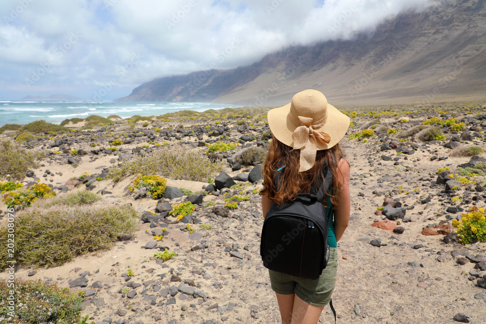 Adventure travel concept. Young woman looking to impressive fantastic landscape of Lanzarote. Back view of female figure with straw hat walking and looking to spectacular sight on Lanzarote Island. 