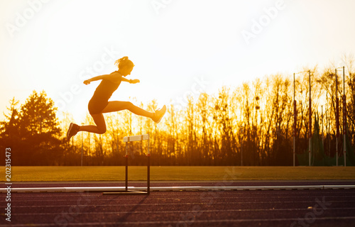 Athletic woman jumping above the hurdle on stadium running track during sunset photo