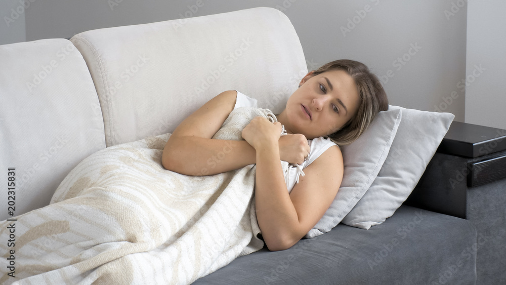 Young ill brunette woman feeling cold while lying under blanket on sofa Young ill brunette woman feeling cold while lying under blanket on sofa