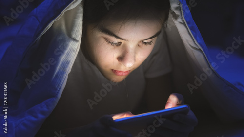 Closeup portrait of girl lying under blanket and playing on mobile phone