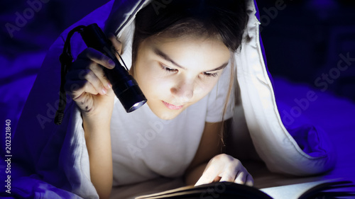 Portrait of teenage girl under blanket with flashlight reading book