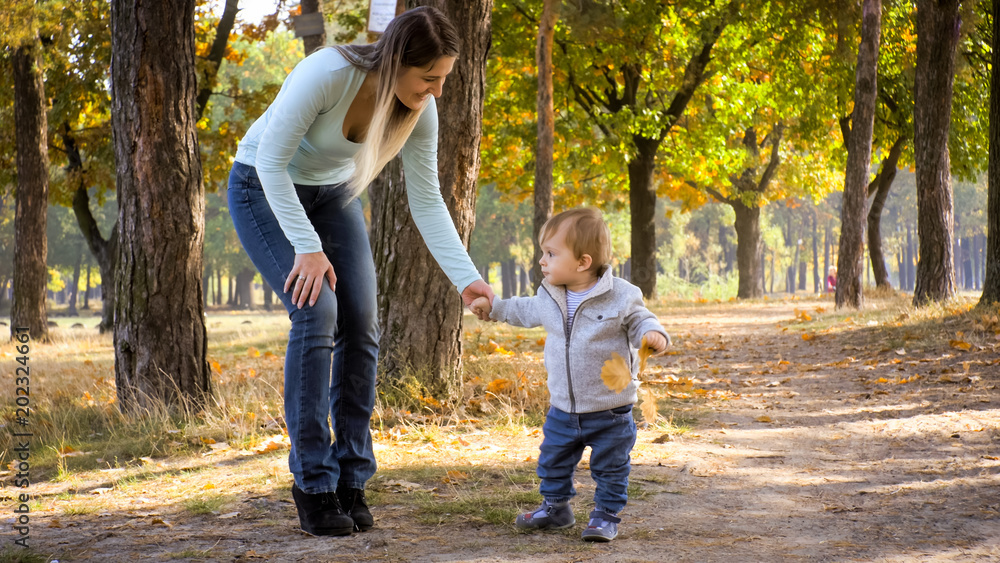 Young smiling mother holding her son by hand while walking at autumn park