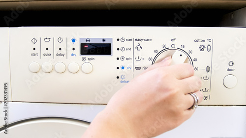 Closeup image of young woman turning round knob on washing maching control panel and choosing water temperature