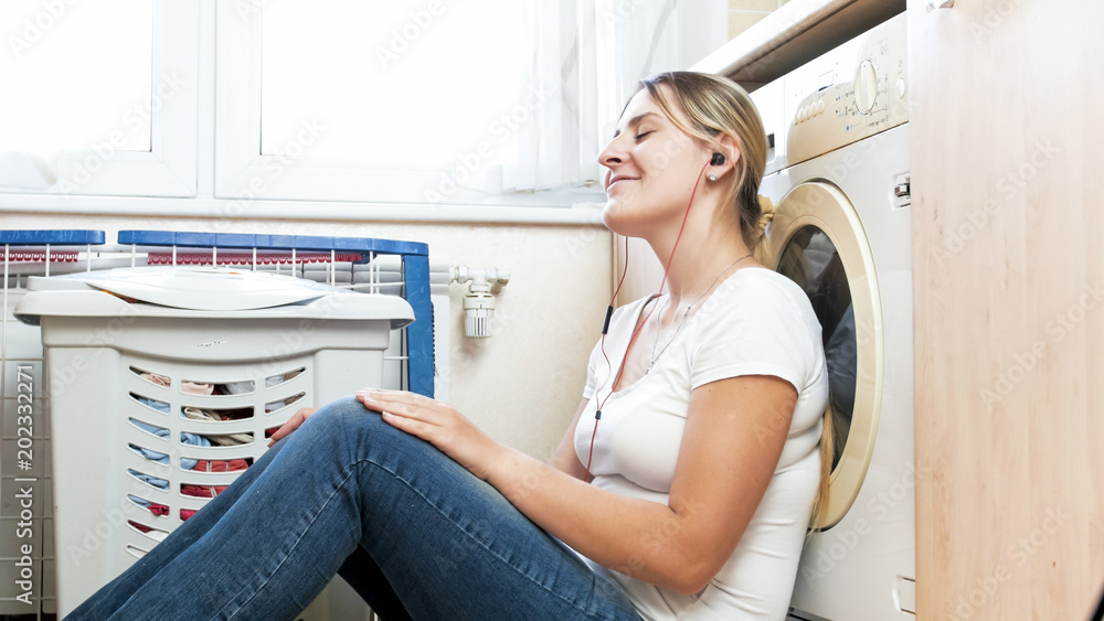 Beautiful smiling woman sitting on floor at laundry room and listening to music Beautiful smiling woman sitting on floor at laundry room and listening to music