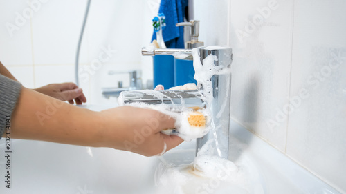 Closeup photo of young woman applying detergent on yellow sponge to water tap and sink