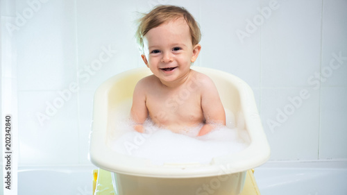 Happy smiling toddler boy sitting in small plastic bathtub with suds
