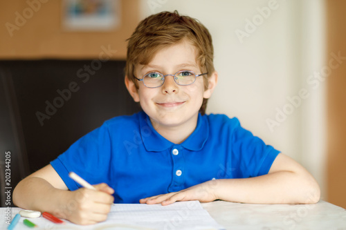 Cute little kid boy with glasses at home making homework, writing letters with colorful pens.