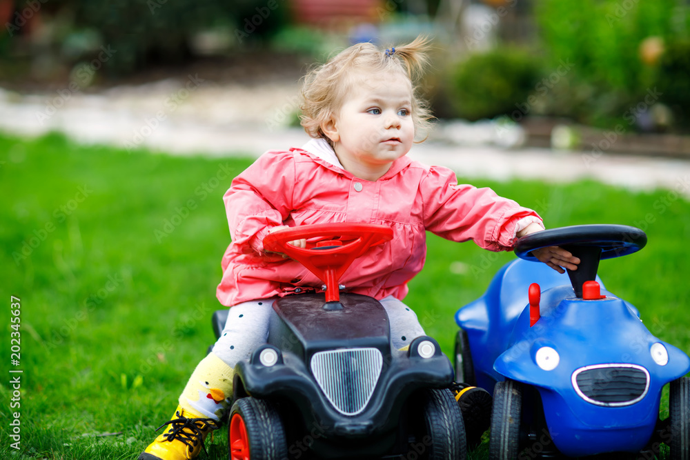 Cute little baby girl playing with two toy cars in garden. Adorable toddler child having fun. Girl in colorful fashion clothes. Cute little baby girl playing with two toy cars in garden. Adorable toddler child having fun. Girl in colorful fashion clothes.
