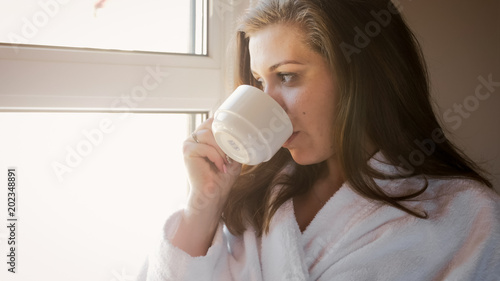 Closeup portrait of young woman looking out of the window and drinking coffee