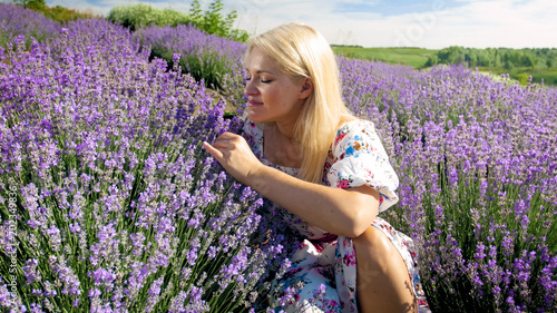 Beautiful young woman in dress sitting in lavender field and smelling flowers