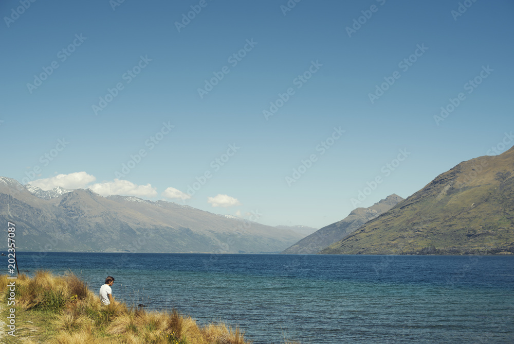 Joven sentado frente a un gran lago azul con montañas de fondo en un día luminoso de verano