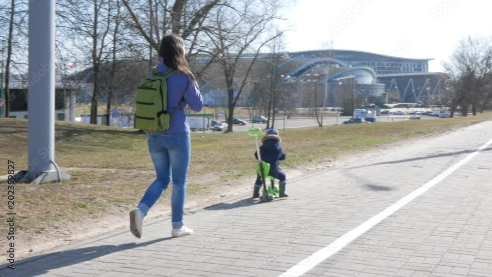 A young mother and a boy are walking outside. The kid skates sitting on the scooter A young mother and a boy are walking outside. The kid skates sitting on the scooter