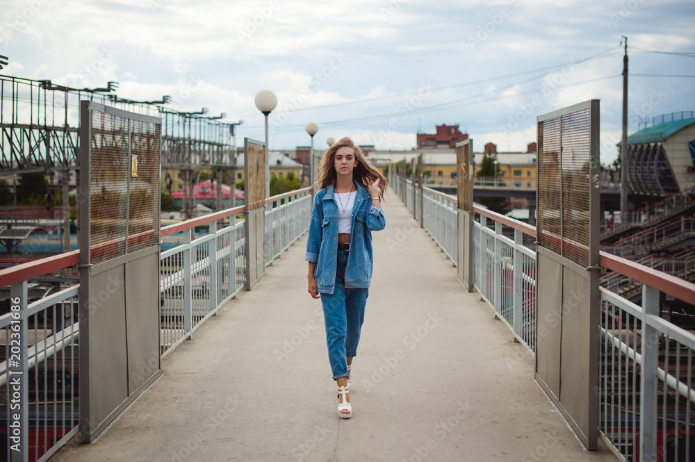 street portrait of a young attractive emotional girl with curly slips dressed in a trendy blue jeans suit on a style walking outdoors against the backdrop of a train station