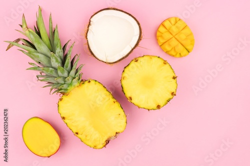 Tropical fruit flat lay with pineapple, mango, and coconut on a pastel pink background. Corner orientation.