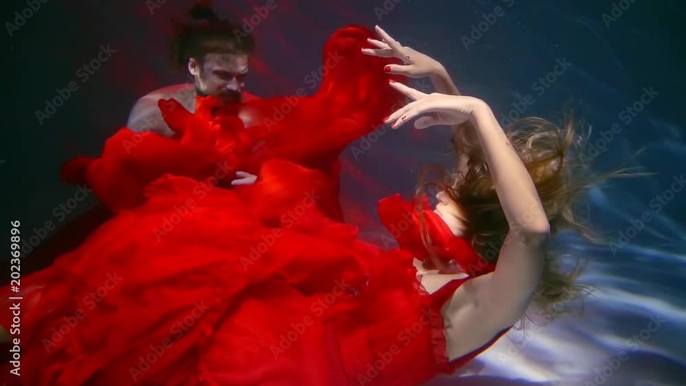 a woman dressed in a red dress swims under the water along with a man, her dress fluttering while bathing a woman dressed in a red dress swims under the water along with a man, her dress fluttering while bathing