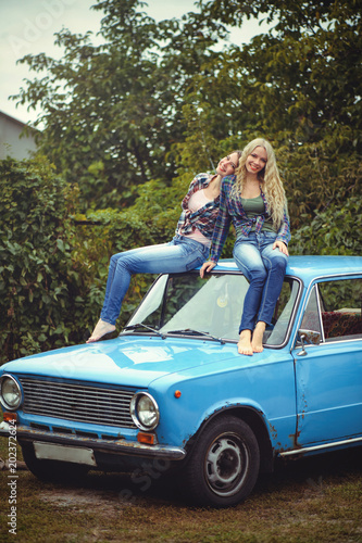 cheerful Attractive two young blonde girl and brunette posing on the hood of an old rusty car, dressed in jeans and shirts on a nature background