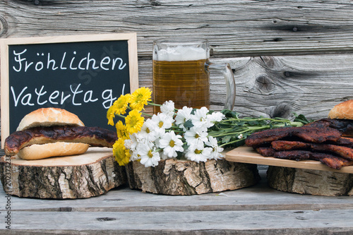 Text "Fröhlichen Vatertag" mit Bratwurst und Bauchfleisch  und Bier rustikal auf Holz
