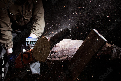 Photo of a worker in outfit with blue gloves cutting wood log on black background.