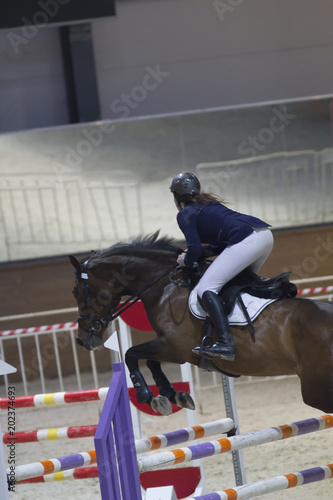Young woman on the black stallion jumping over hurdle at show jumping competition