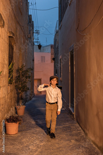 a young village worker walks through the narrow ancient streets of the city in an old-fashioned simple clothes with a guitar on his shoulder photo