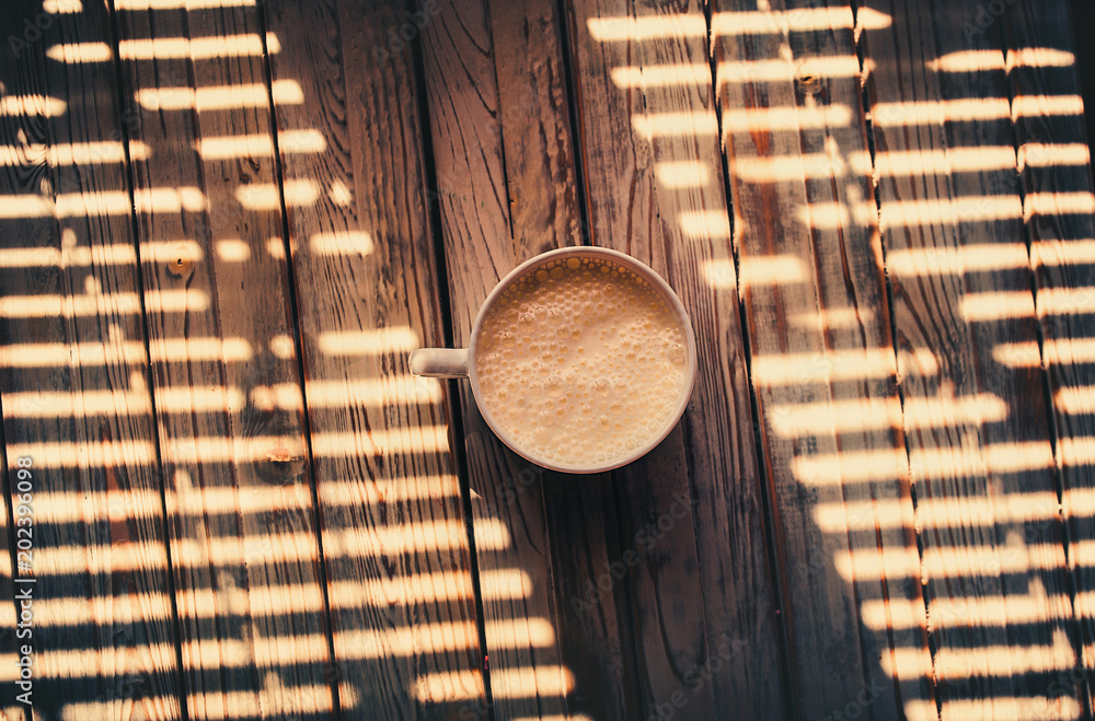Φωτογραφία White cup with yogurt stands on a wooden background in the shade of the blinds in the light of the bright sun