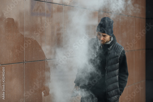 Vape teenager. Portrait of a handsome young white guy in black waistcoat and modern cap vaping an electronic cigarette opposite the futuristic modern background. Lifestyle.