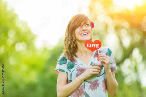 Cute young woman holding a paper heart - shaped sign outdoors.