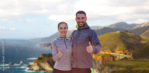 fitness, sport and gesture concept - smiling couple outdoors showing thumbs up over bixby creek bridge on big sur coast of california background