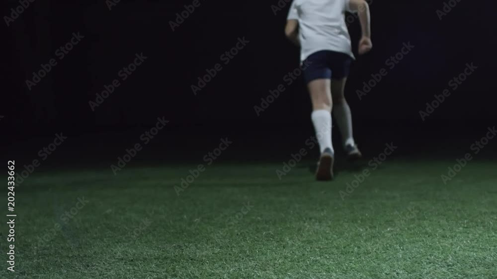 Rear view of young soccer player shooting a ball and running away while training on artificial turf in dark arena Rear view of young soccer player shooting a ball and running away while training on artificial turf in dark arena