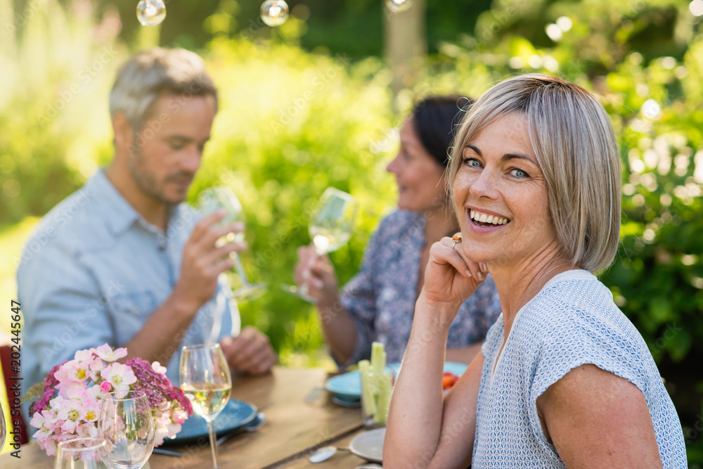 A bunch of friends gathered to share a meal around a table in the garden. Focus on a beautiful woman looking at the camera A bunch of friends gathered to share a meal around a table in the garden. Focus on a beautiful woman looking at the camera