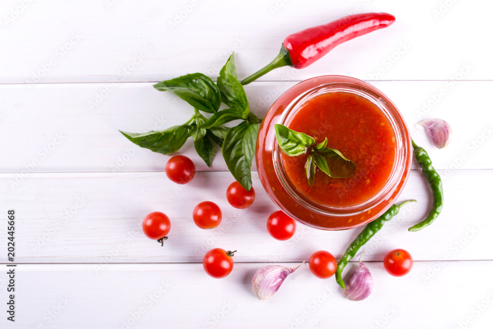 Tomato ketchup sauce with cherry tomatoes and red hot chili peppers, garlic and herbs in a glass jar on white background. Homemade tomato sauce and fresh tomatoes. . Flat lay, top view Tomato ketchup sauce with cherry tomatoes and red hot chili peppers, garlic and herbs in a glass jar on white background. Homemade tomato sauce and fresh tomatoes. . Flat lay, top view