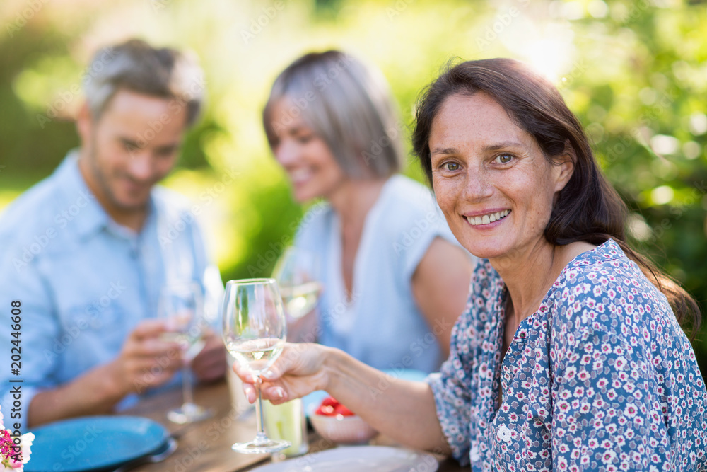 A group of friends gathered to share a meal around a table in the garden. Focus on a beautiful woman looking at the camera A group of friends gathered to share a meal around a table in the garden. Focus on a beautiful woman looking at the camera