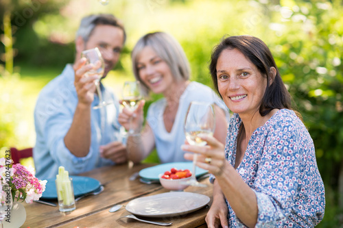 In summer. a group of friends in their forties gathered around a table in the garden to share a meal. They toast with their glasses of wine to the camera.