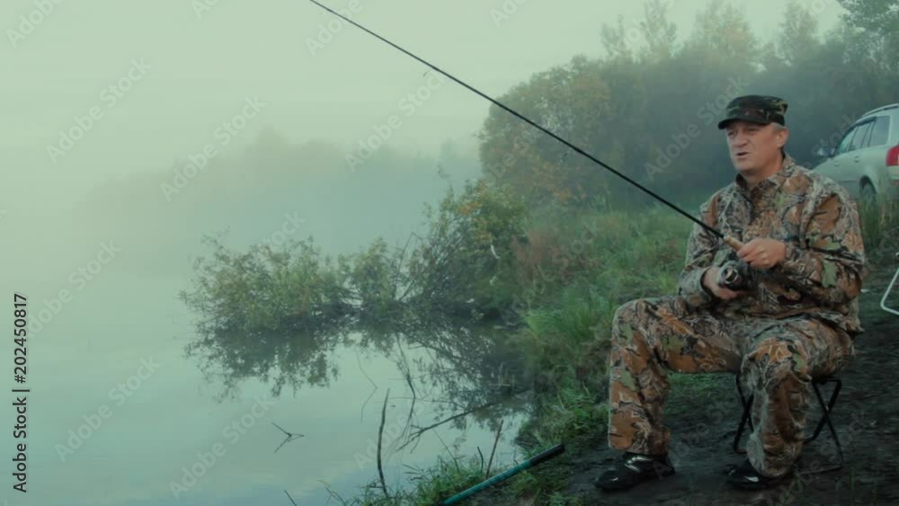 Fisherman with spinning The general plan of the fisherman sitting on a spreading chair on the shore of the lake on a foggy morning with spinning Fisherman with spinning The general plan of the fisherman sitting on a spreading chair on the shore of the lake on a foggy morning with spinning
