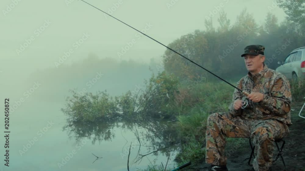Fisherman with spinning The general plan of the fisherman sitting on a spreading chair on the shore of the lake on a foggy morning with spinning Fisherman with spinning The general plan of the fisherman sitting on a spreading chair on the shore of the lake on a foggy morning with spinning