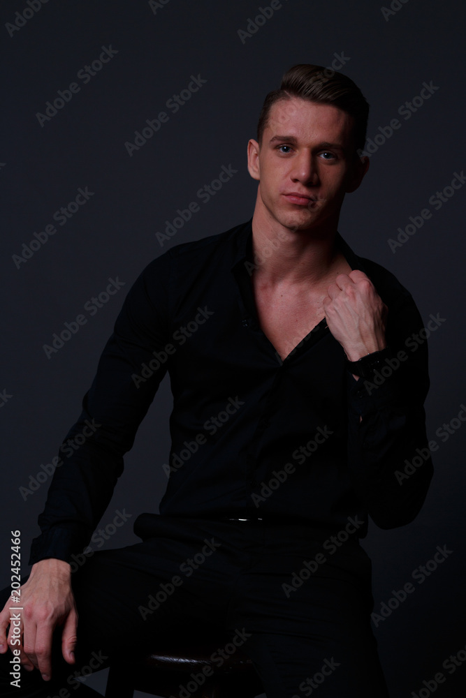 a young, white guy sits on a chair in a black shirt and black pants,  against a black background,straightens the shirt and looking at camera.  business, model, interview, portrait Stock Photo